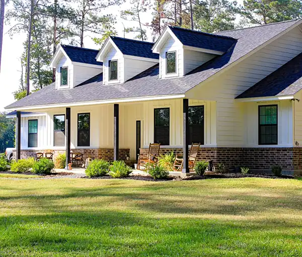 Front porch of a home with three roof dormers, brick skirting and rocking chairs along the covered porch.